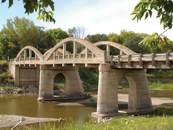 An impressive triple-arch concrete bridge over the Assiniboine River, in the village of Millwood in the Rural Municipality of Russell, was built in 1920, replacing an earlier wooden structure. Aside from minor damage to the balusters of its railings, it is in good condition.