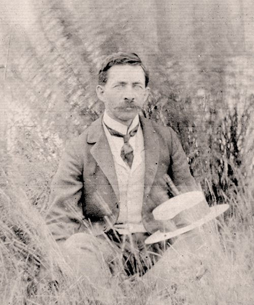 Man with moustache sitting in tall grass Sisler posed for the camera in front of his one-room school at New Stockholm, Saskatchewan Territory in 1900, the place where he first developed his ideas on the ‘direct method’ of teaching English.